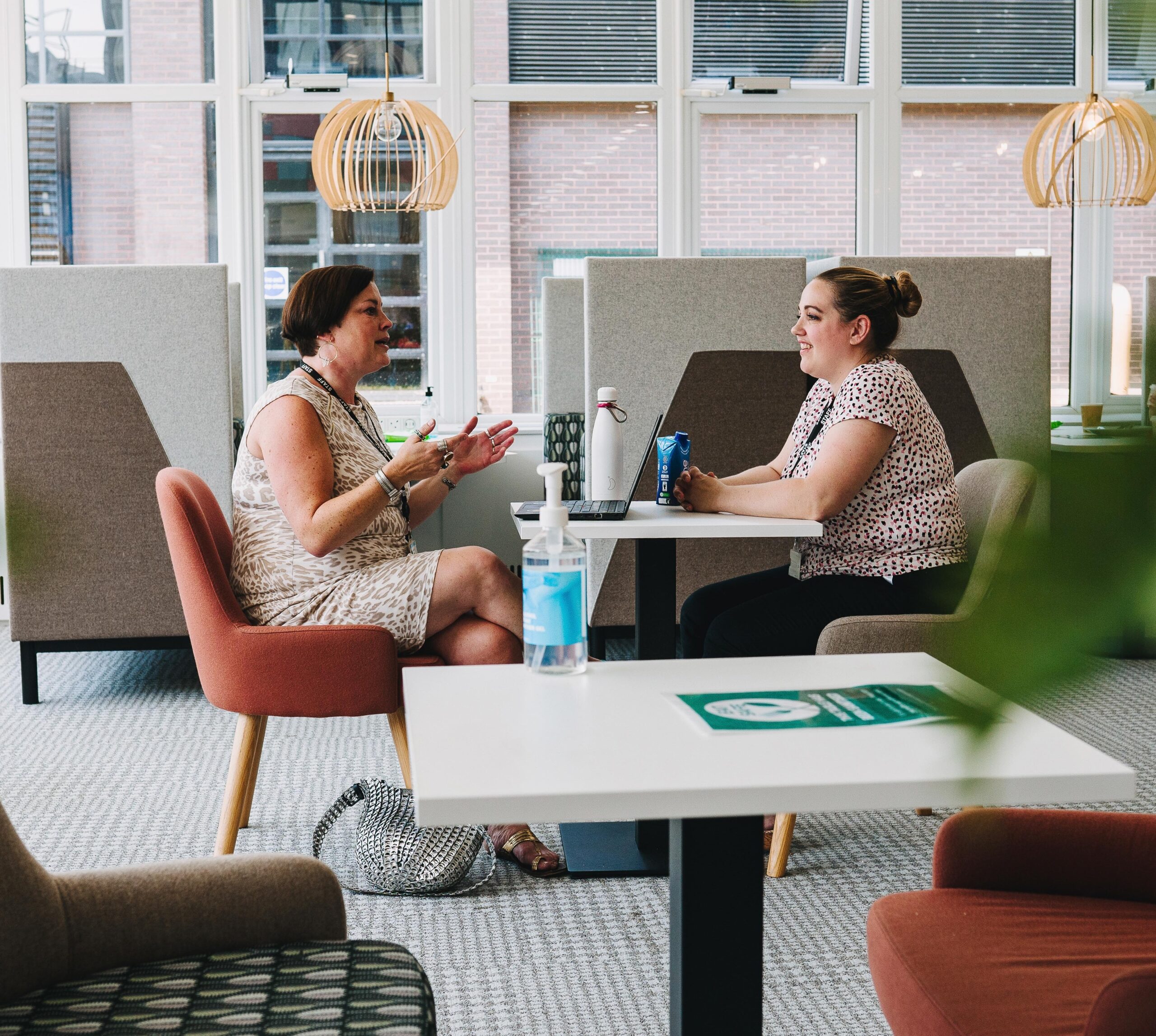 Two women smiling engaged in conversation in one of the relaxed the ONS cafe seating meeting areas. They are sat facing each other at a white table in modern upholstered chairs. One woman gestures with her laptop open in front of her while sat opposite the other woman smiles.