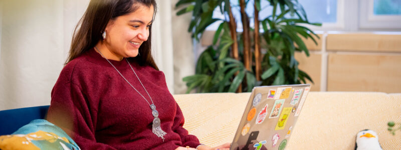 A woman smiles as she sits on a sofa with a laptop in her living room. She appears to be typing.