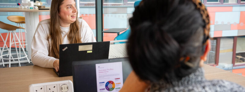 Two women sit opposite each other in a glass walled office, one with her face to the camera the other faces away. They review the civil service success profiles and appear engaged in discussion.